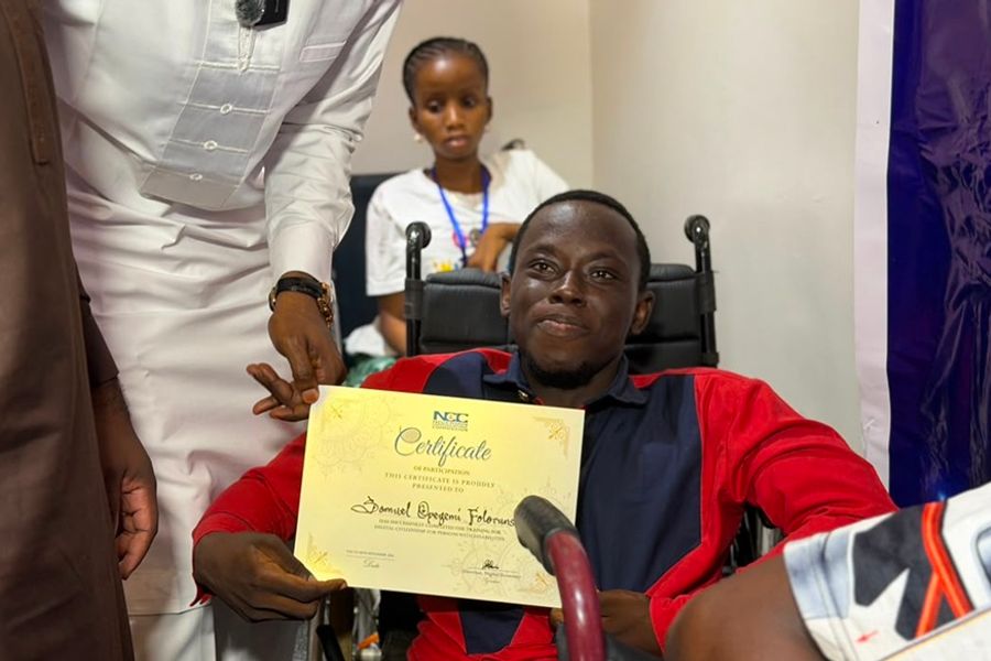 A smiling participant seated in a wheelchair holds up a certificate presented at the end of a digital citizenship training programme organised for Persons with Disabilities. Other participants and event representatives are visible in the background and foreground. The certificate bears the Nigerian Communications Commission (NCC) logo and recognises the participant’s completion of the programme.